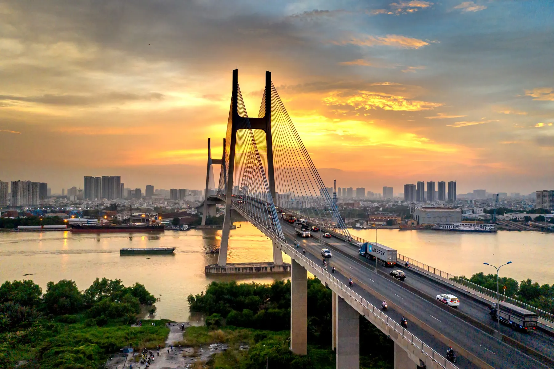 A cable-stayed bridge with two towers across a river at sunset, the city behind